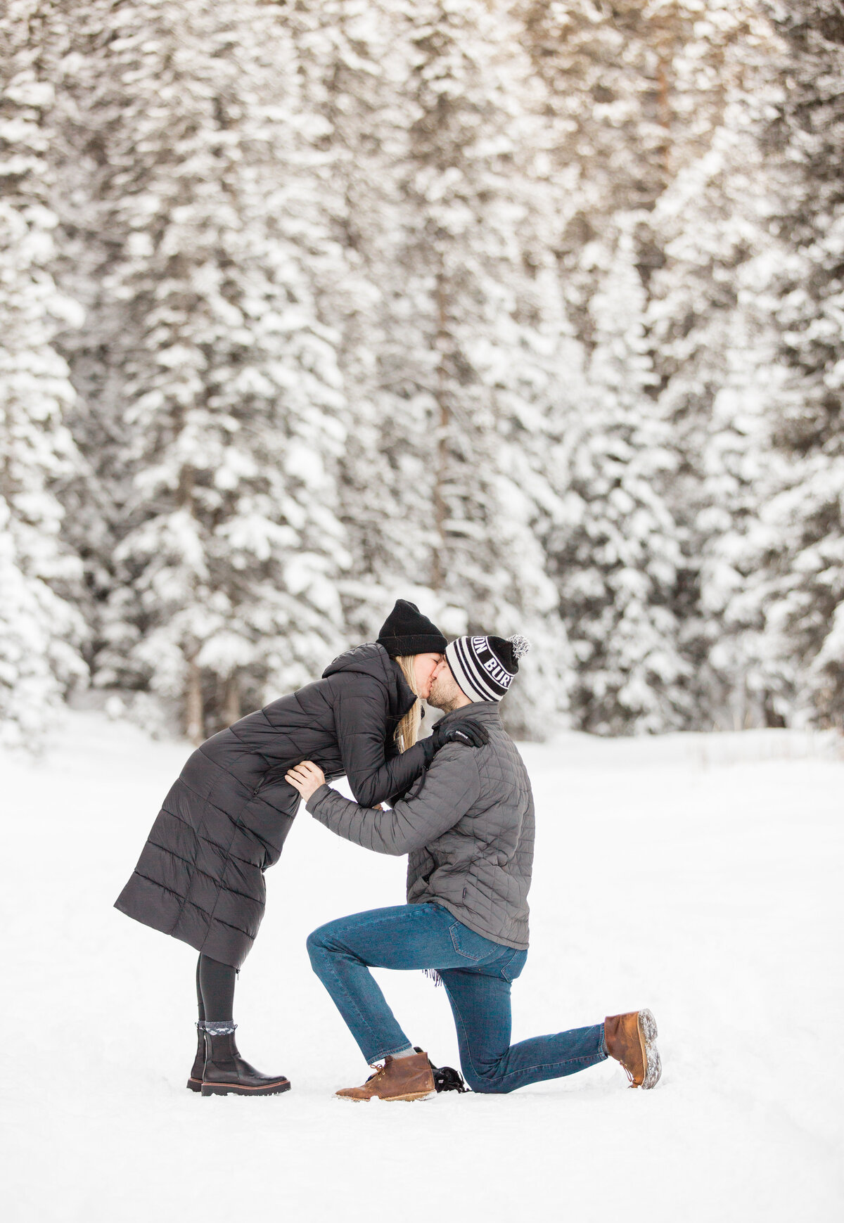 A woman knees down to kiss her fiance who has just asked her to marry him. They are in a gorgeous winter scene with snow covered pine trees