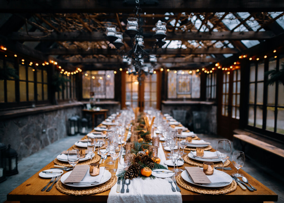 A large wooden table is set in a glasshouse with oranges and pinecones for a dinner party.