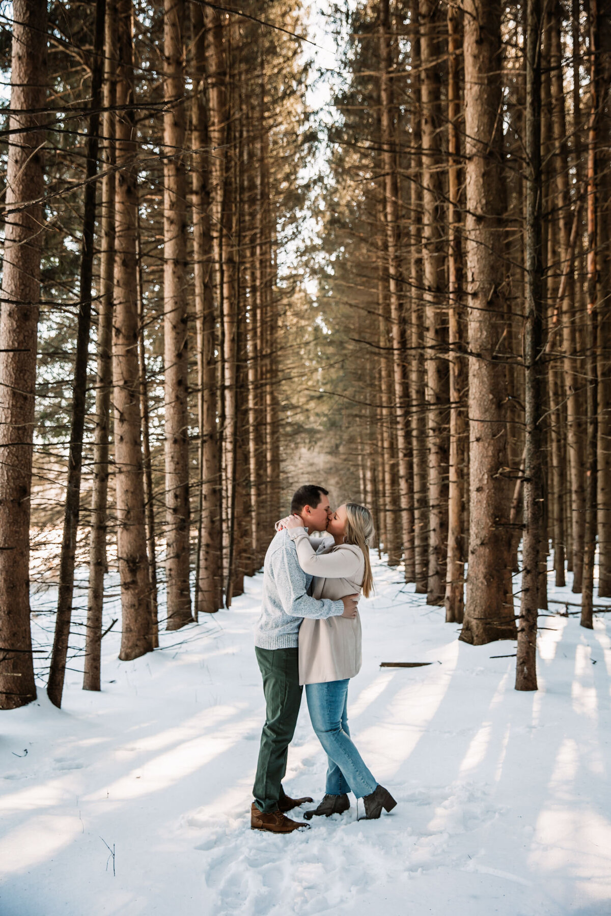 man and woman kiss in a row of trees in the middle of a forest during winter