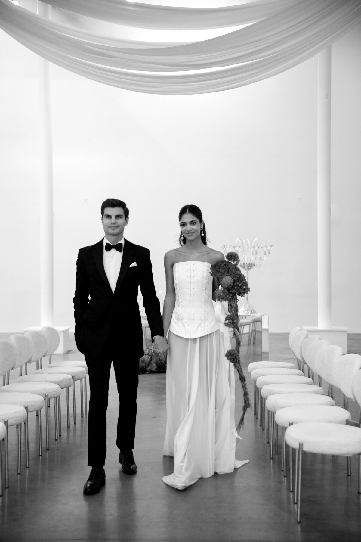 Black and white portrait of a bride and groom seated together at a modern wedding table inside a minimalist NYC studio.