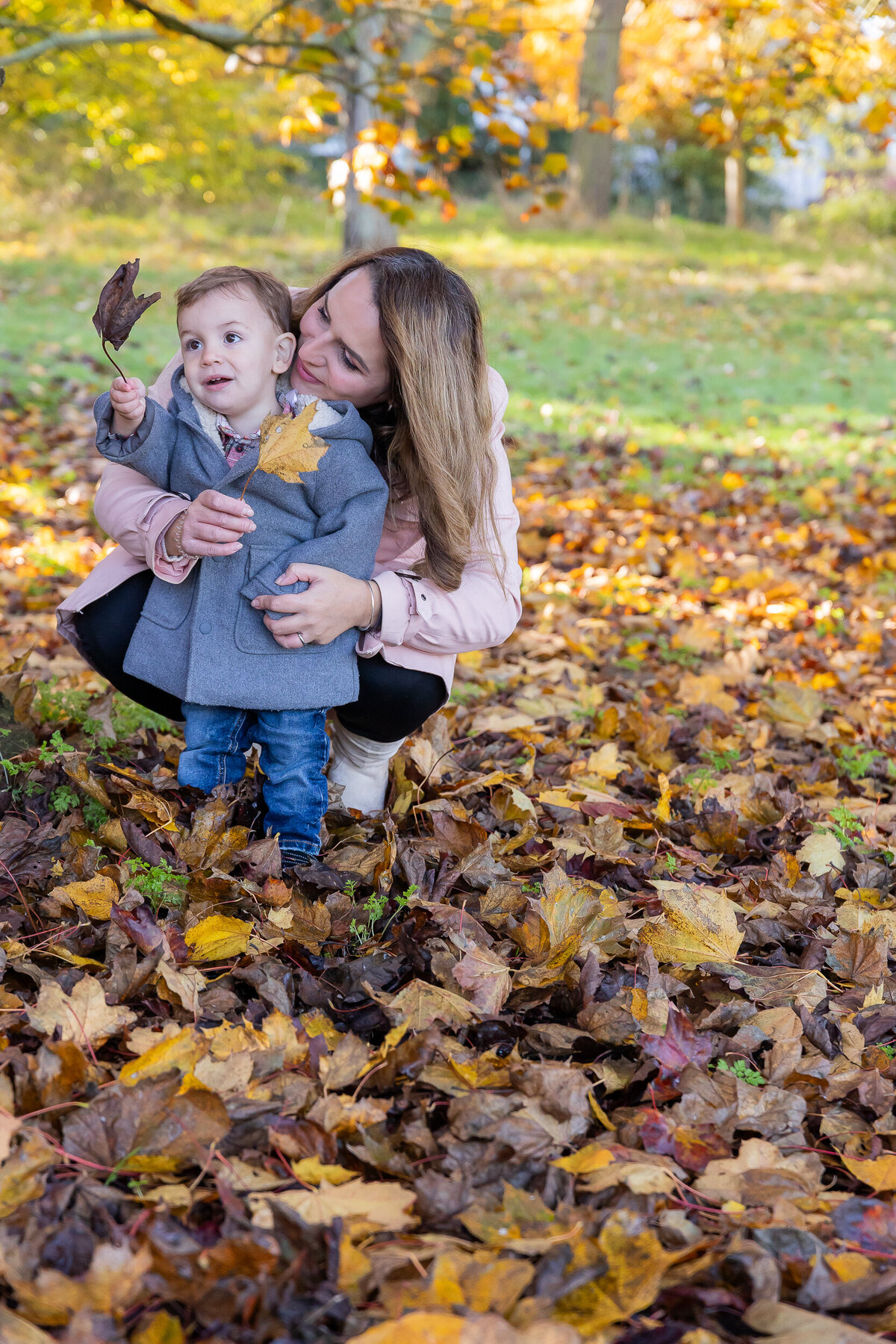 A mother and her young son joyfully holding autumn leaves in a park, surrounded by fallen leaves on the ground.