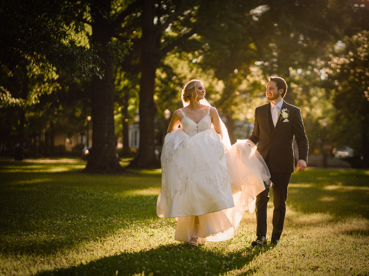 A groom holding a bride's train as they walk in a field.