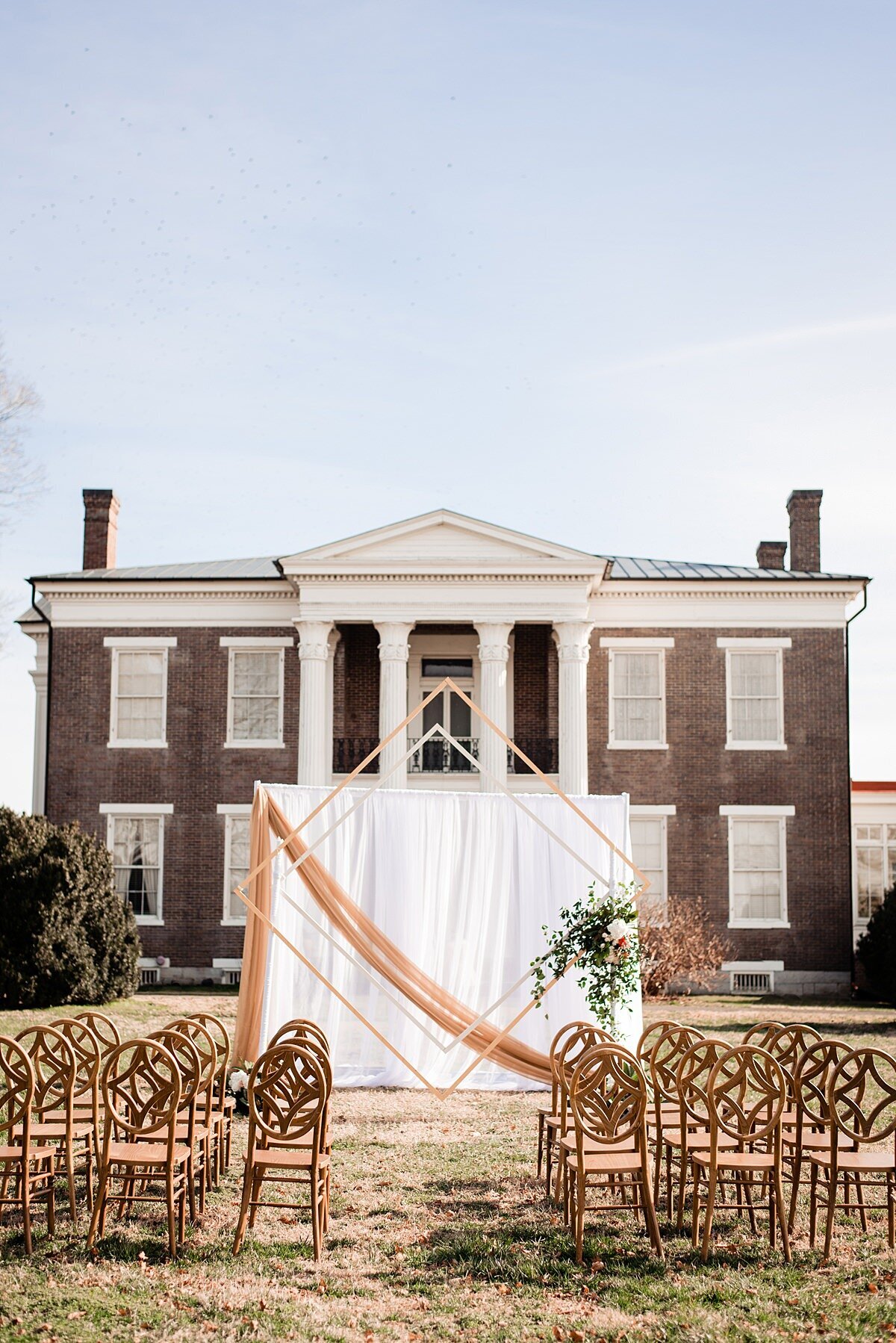Art deco ceremony arbor with drapery and wood banquet chairs set on the front lawn in front of Rippa Villa mansion