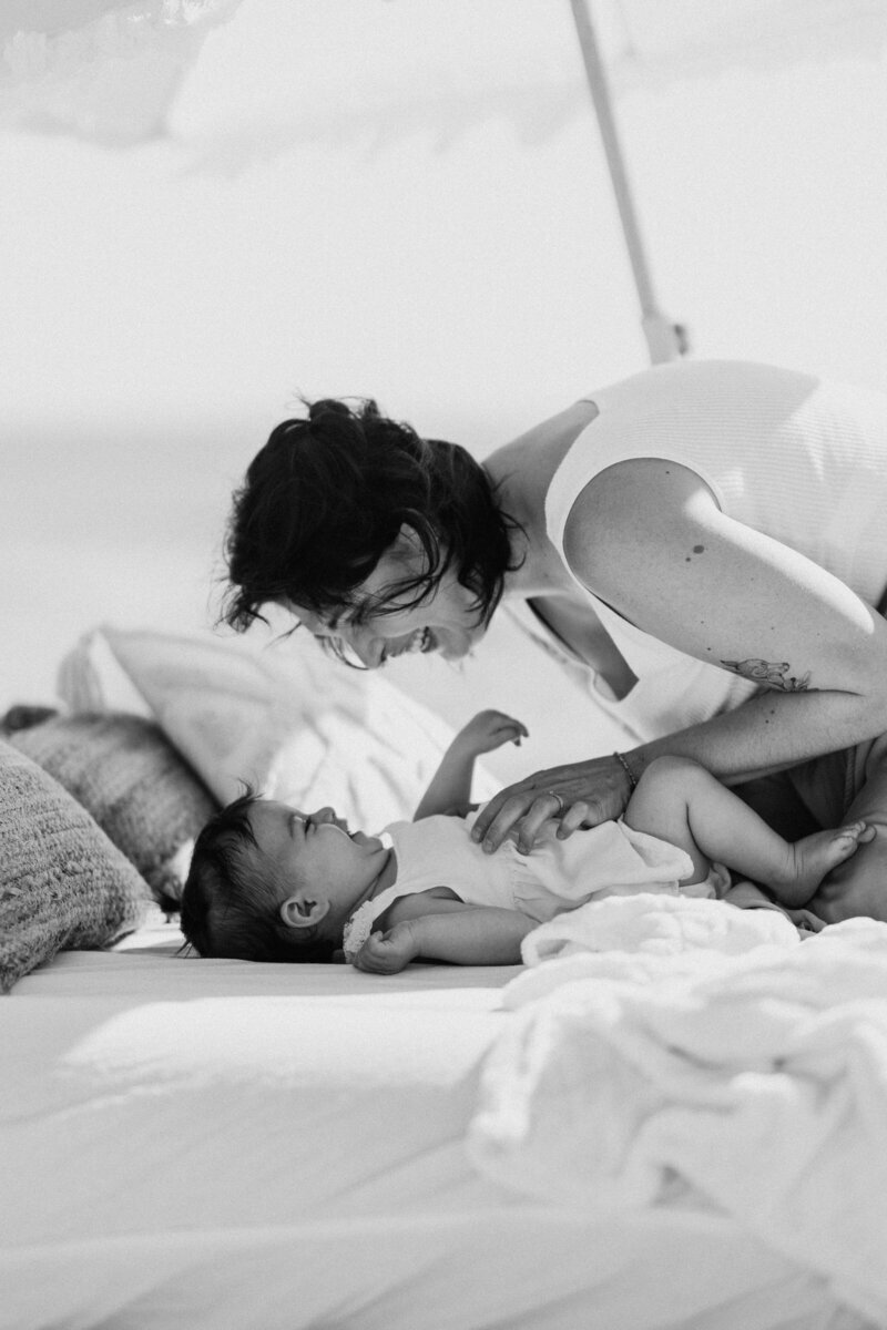 Maman faisant des chatouilles à sa fille allongée dans un décor cosy sous une ombrelle à la plage lors d'une séance photo en Vendée.