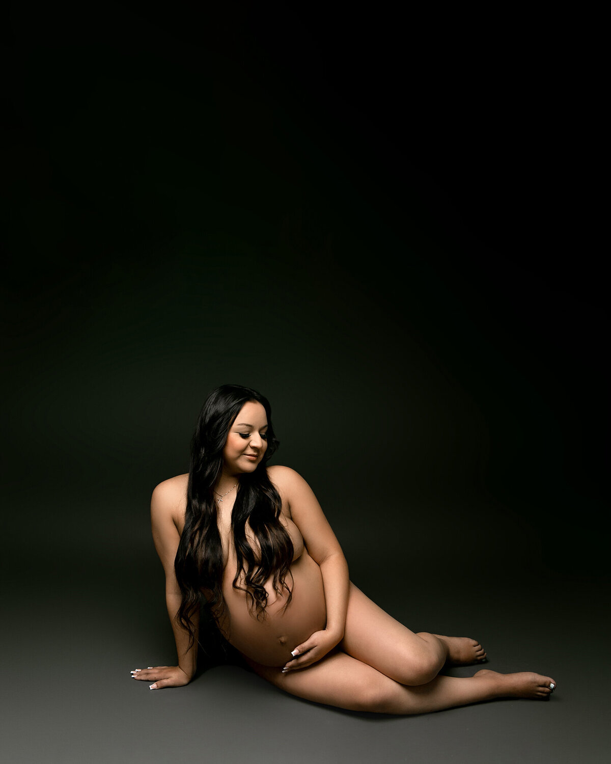 Expectant woman sitting on the ground unclothed holding her pregnant belly in a session with Fresno, CA matenity photographer