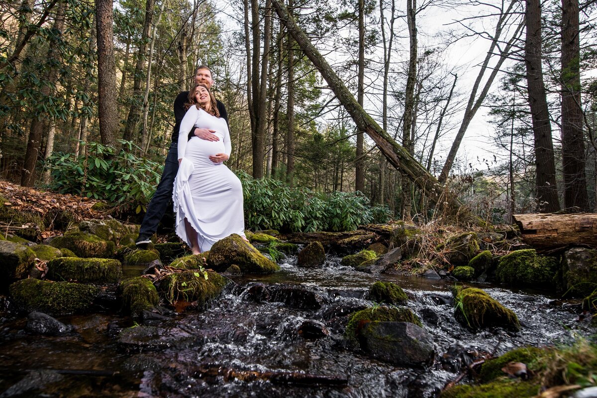 a woman wearing a white maternity dress leans against her partner in front of a babbling brook