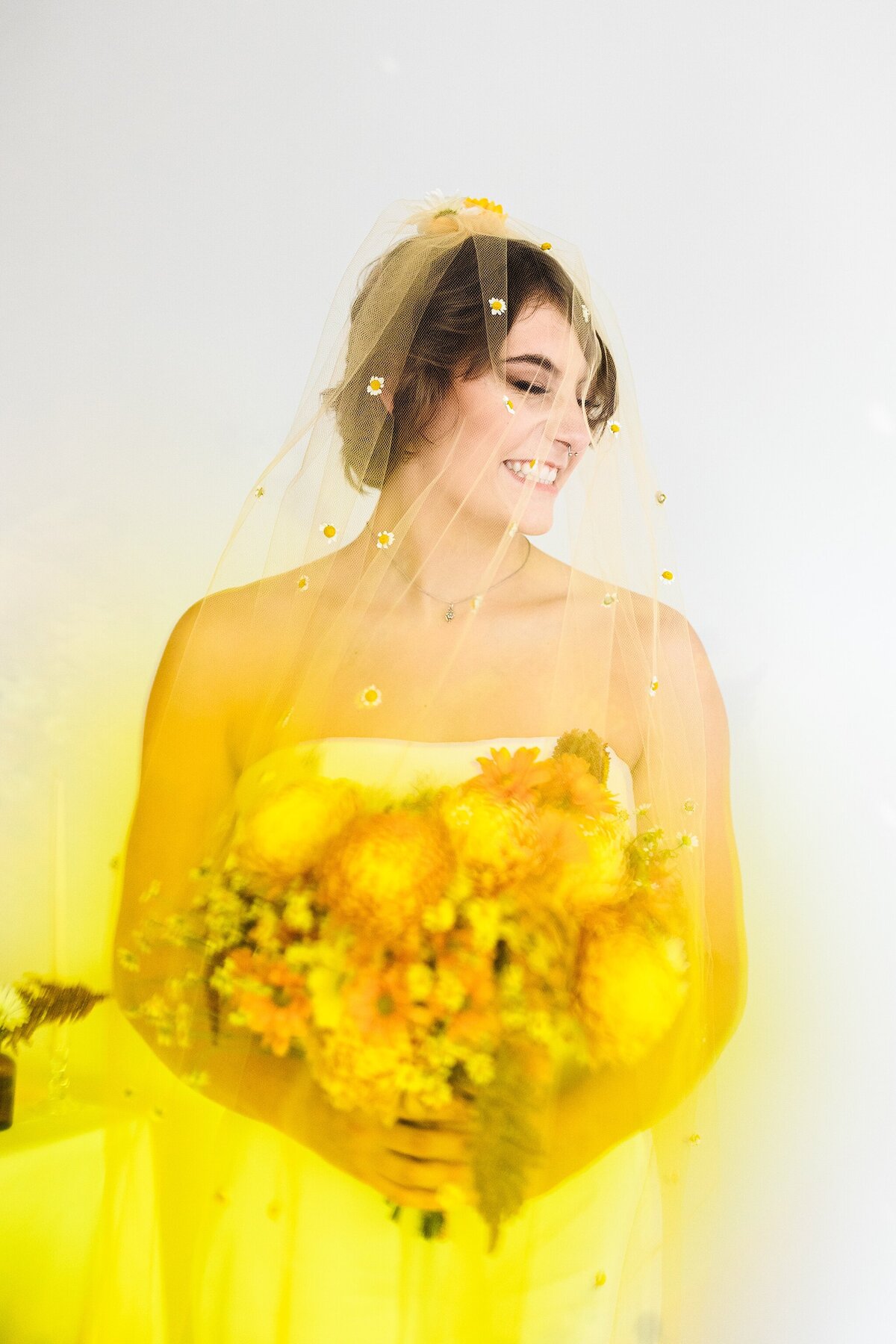 A portrait of a bride posing coyly on her wedding day in DFW, Texas. The bride is wearing a sleeveless white dress with a veil and is holding a bouquet in front of a white background. However, due to a colorful cube being held up in front of the camera lens, the dress (and overall ambiance) appears to be yellow.