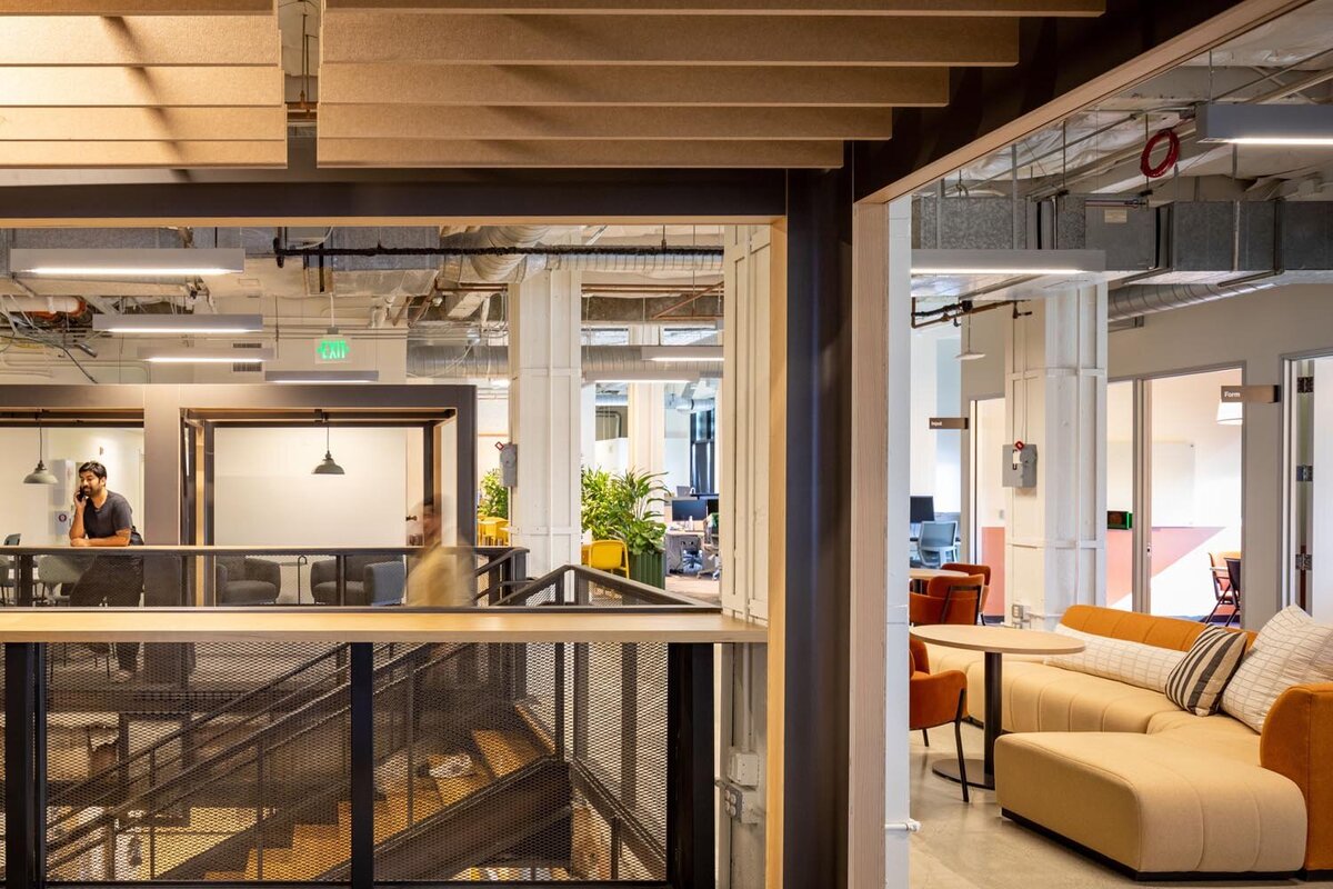 Upper-level view of an open office with acoustic ceiling panels, private meeting pods, lounge seating, and exposed industrial ductwork.