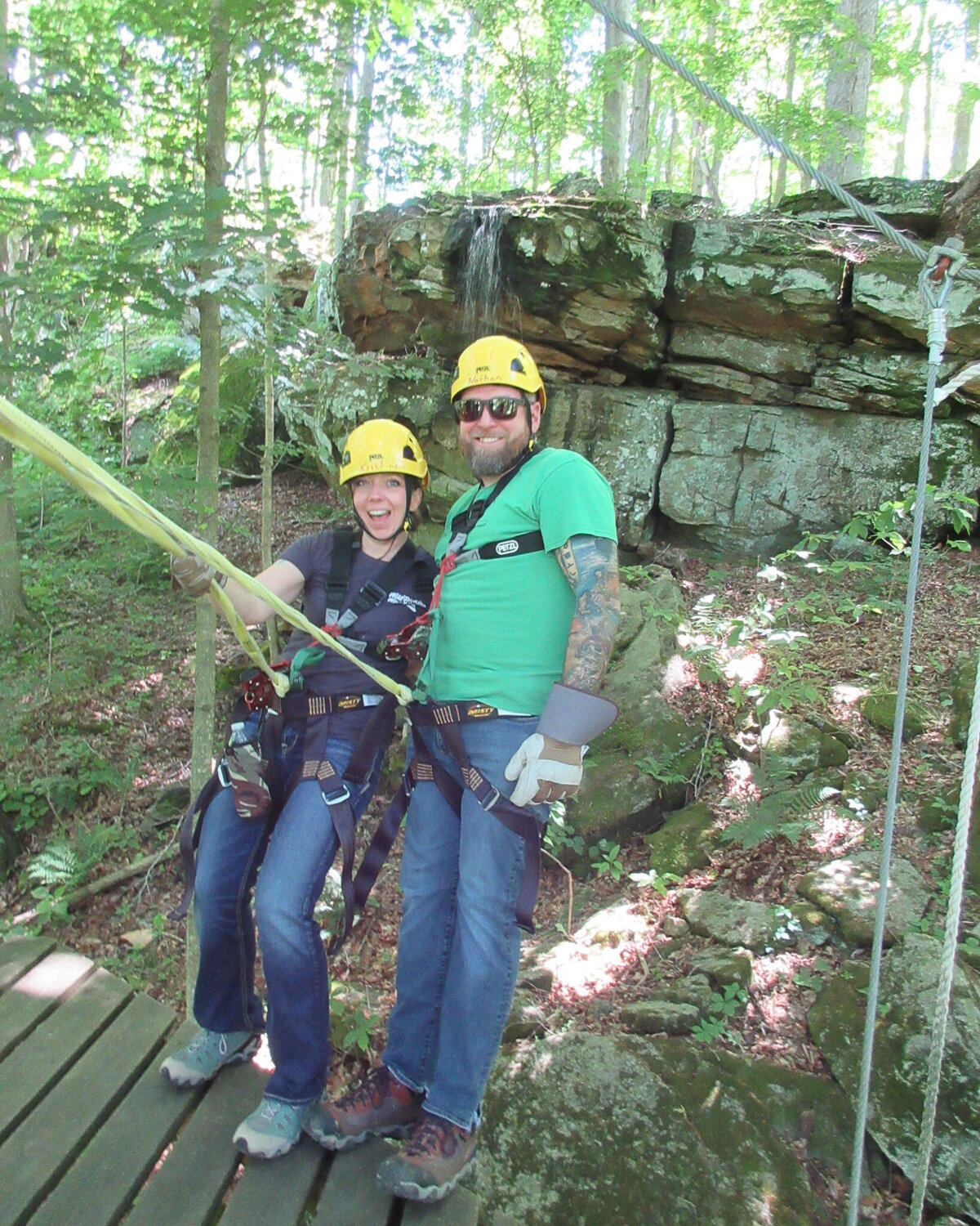 husband and wife zip-lining, wearing hard hats, standing on a bridge with trees in the background smiling at the camera