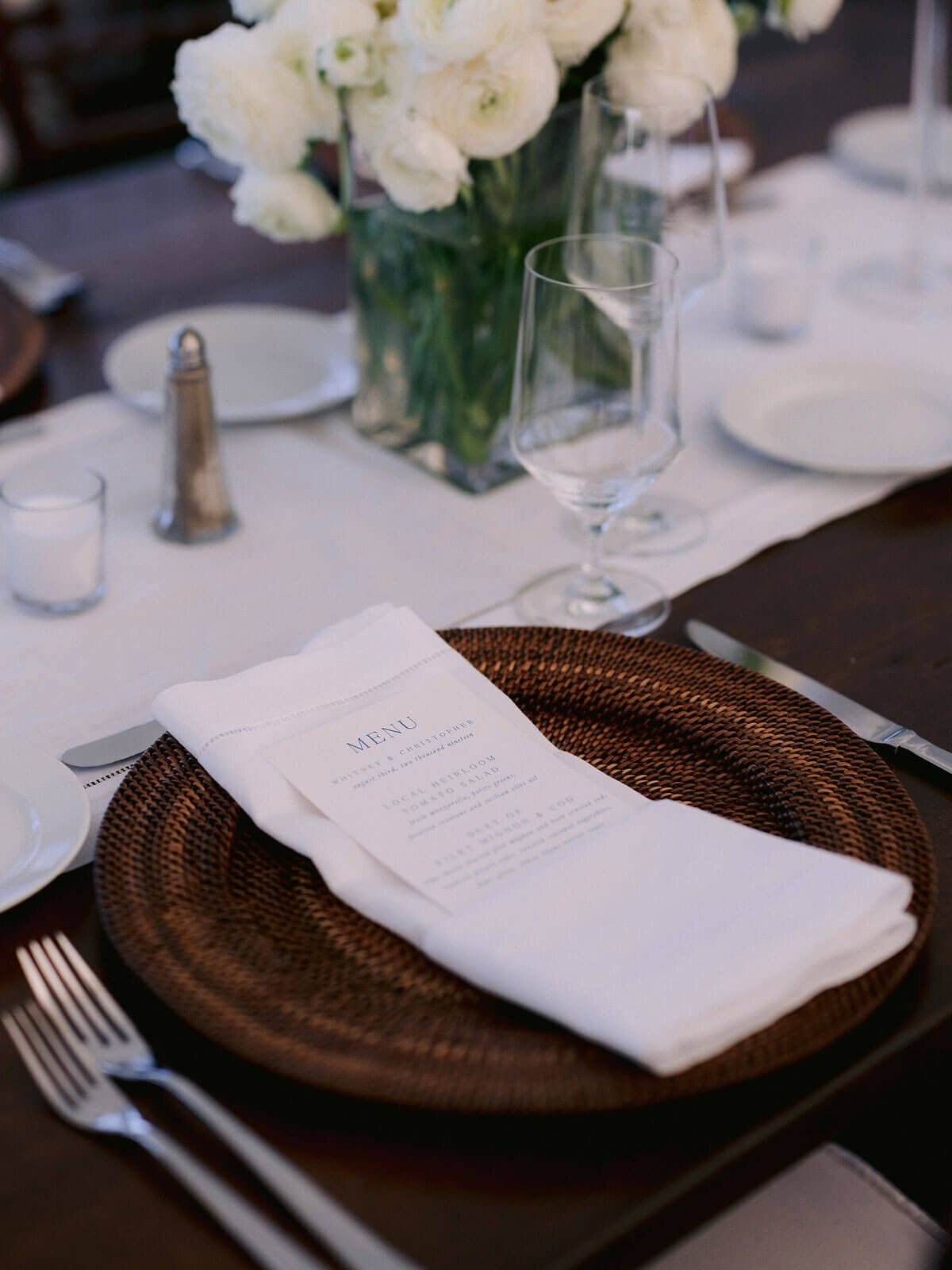 A wicker plate holder, table napkin and Menu, cutleries, glasses, and flowers on a dining table at Cape Cod Summer Tent, MA.