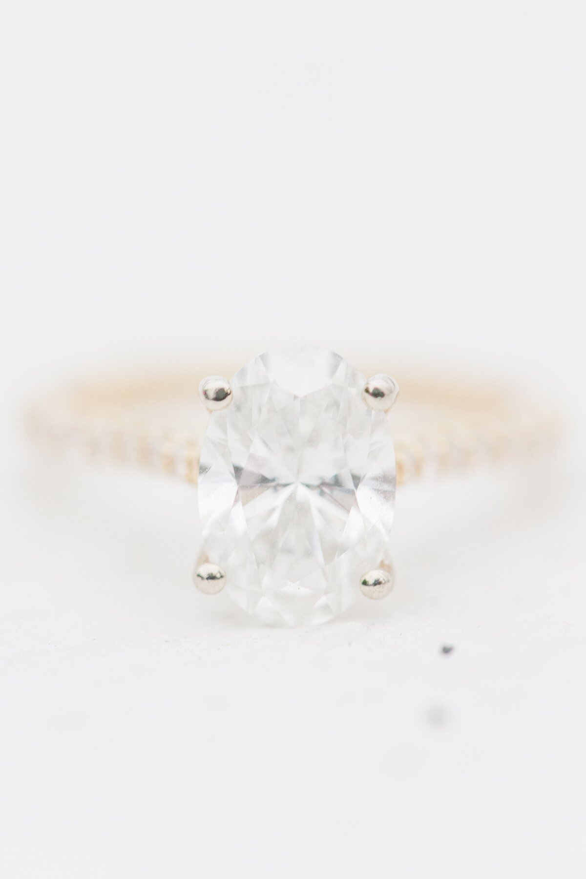 Macro shot of an oval-cut diamond engagement ring with a pavé band against a bright white background — representing the portfolio of a Cape Cod engagement photographer.