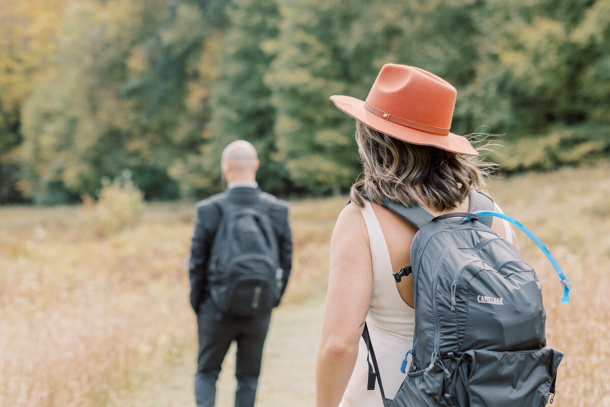 Couple hiking in their wedding clothes. The bride is wearing a burnt orange fedora and a camelbak while the groom is wearing a black suit and backback hiking down the mountain.