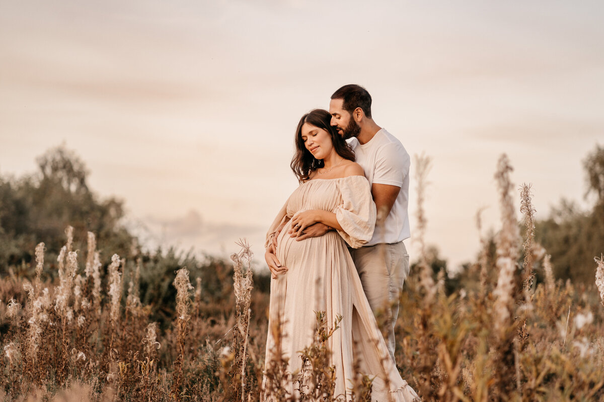 Photo of a pregnant woman and her husband standing outside in a field of long folliage at sunset
