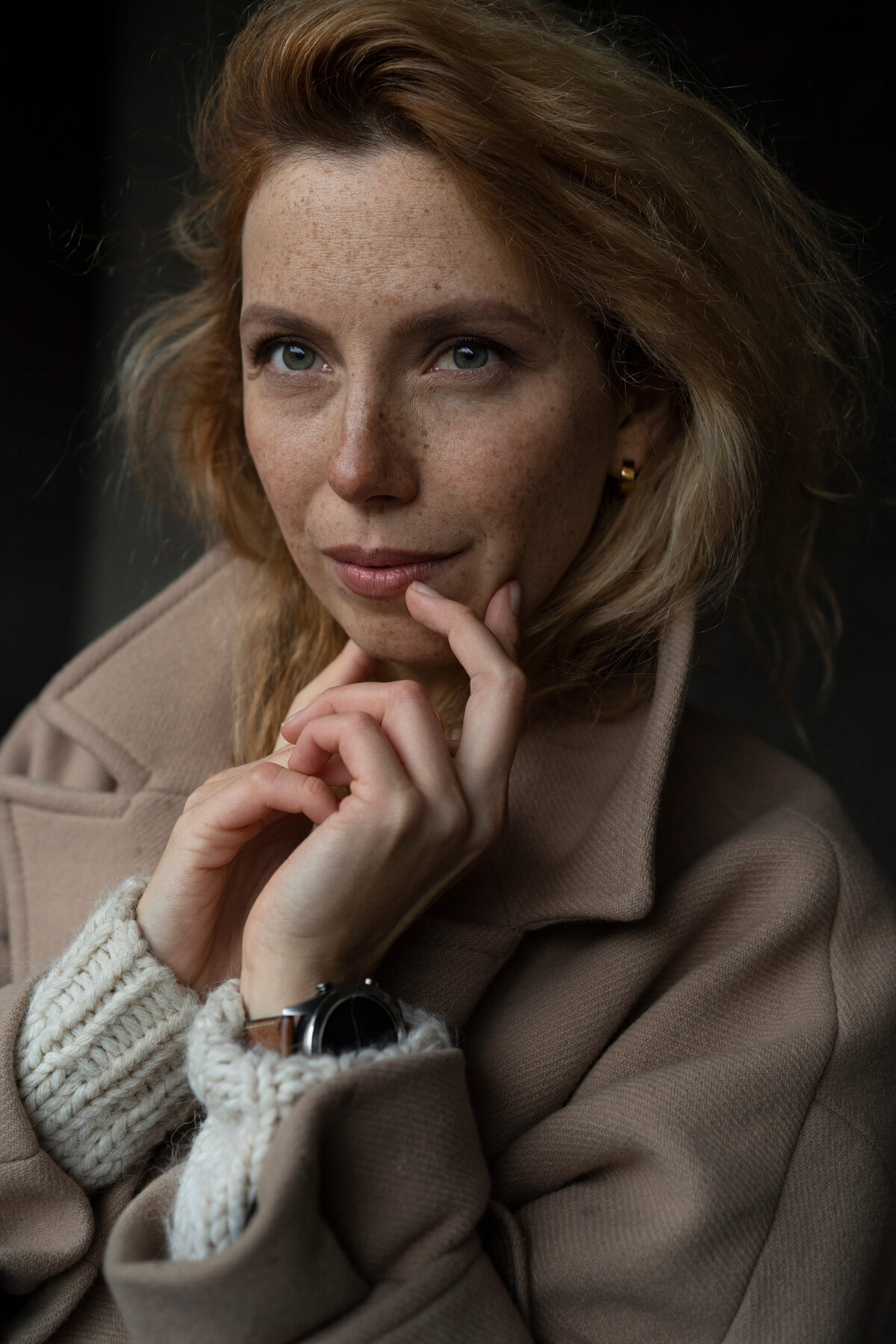 a blond woman with freckles in a beige coat holding her hands by the face