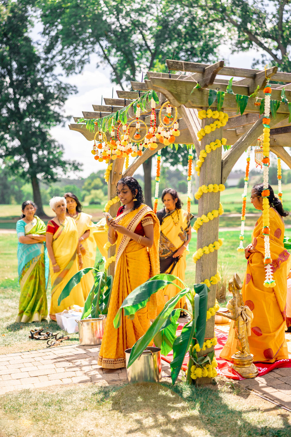 A group of women in colorful traditional sarees stand under a decorated wooden canopy outdoors. The canopy is adorned with yellow flowers and green leaves. One woman is speaking, and there are brass pots and banana leaves around them.