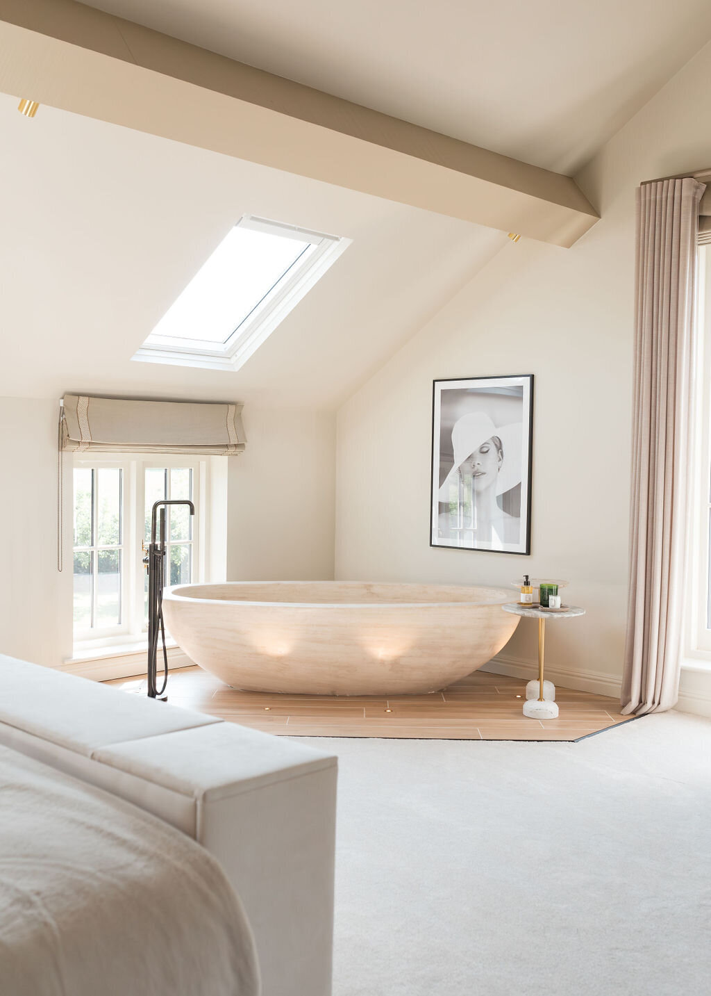 A minimalist neutral bedroom features a large, freestanding wooden bathtub under a skylight. There's a floor-mounted faucet beside the tub, a small side table with decor, and a framed black-and-white photo on the wall. The space is bright with natural light and neutral tones.