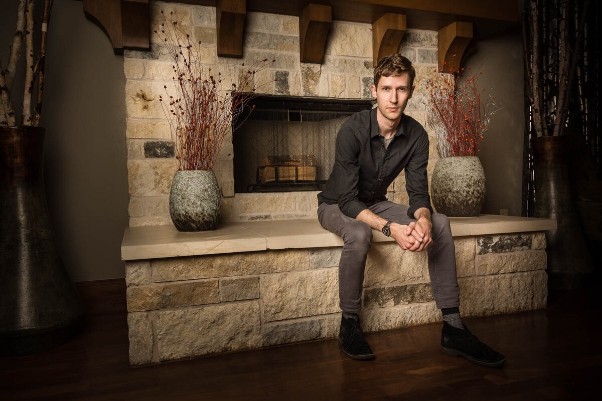 Editorial portrait of a man seated at the base of a stone fireplace in a relaxed, stylish pose.