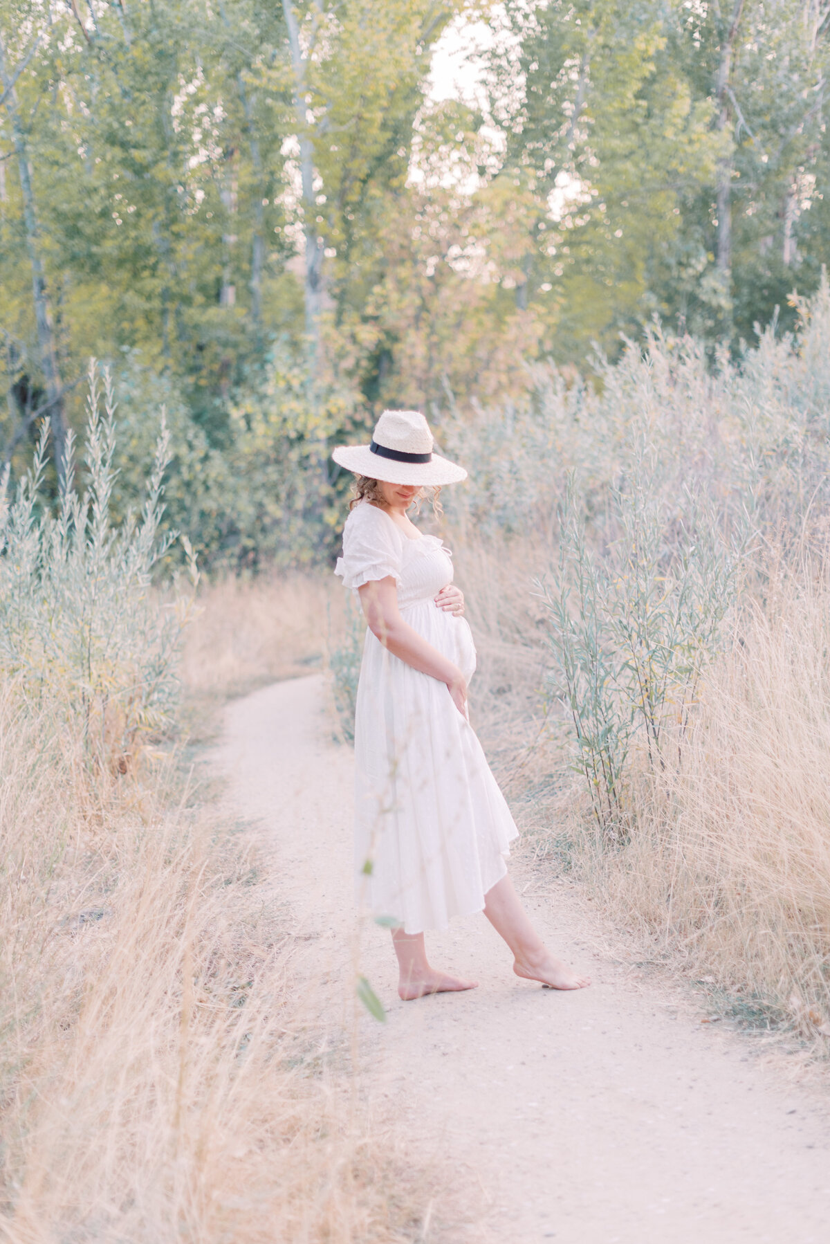 Portrait of a barefoot pregnant woman in a white summer dress and a white hat with her hands around her belly in the country