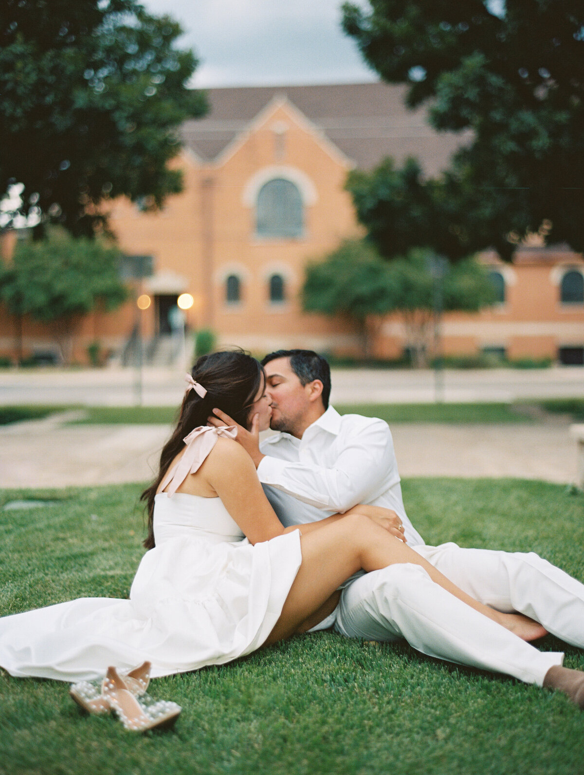 Woman in a white dress kissing a man in a white shirt while sitting in the grass