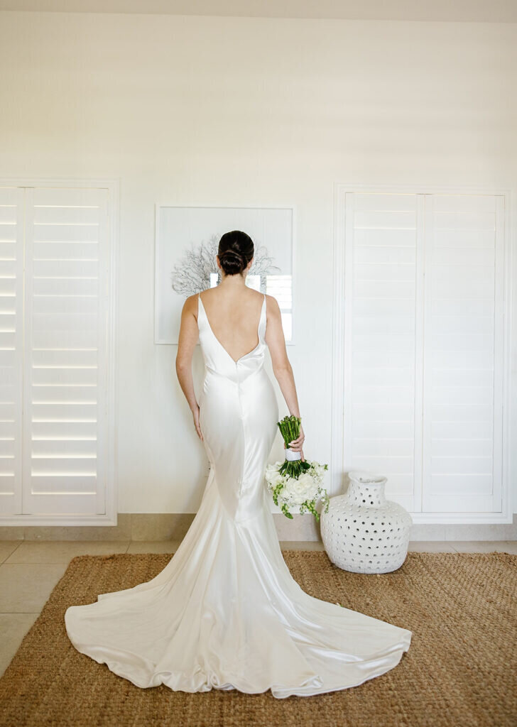 A breathtaking shot of the bride from behind, highlighting the open-back design of her silk wedding dress as she holds a fresh floral bouquet. Destination wedding photography by Claudia Amalia Photography in the Florida Keys.