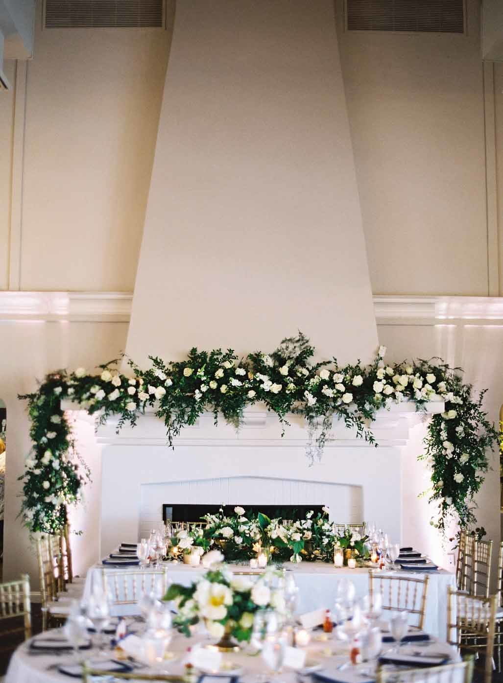 wedding reception head table  in front of large white fire place with greenery arch on top of fireplace mantle