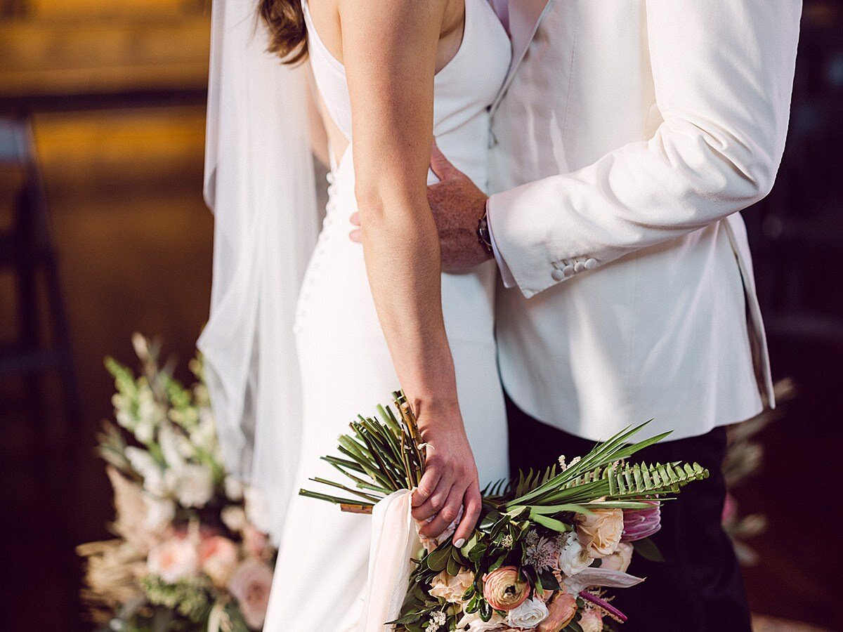 The groom, wearing a tuxedo with a white jacket, embraces the bride who is wearing a silk sheath dress and veil  and is holding a large tropical floral bouquet accented by blush raw silk ribbons