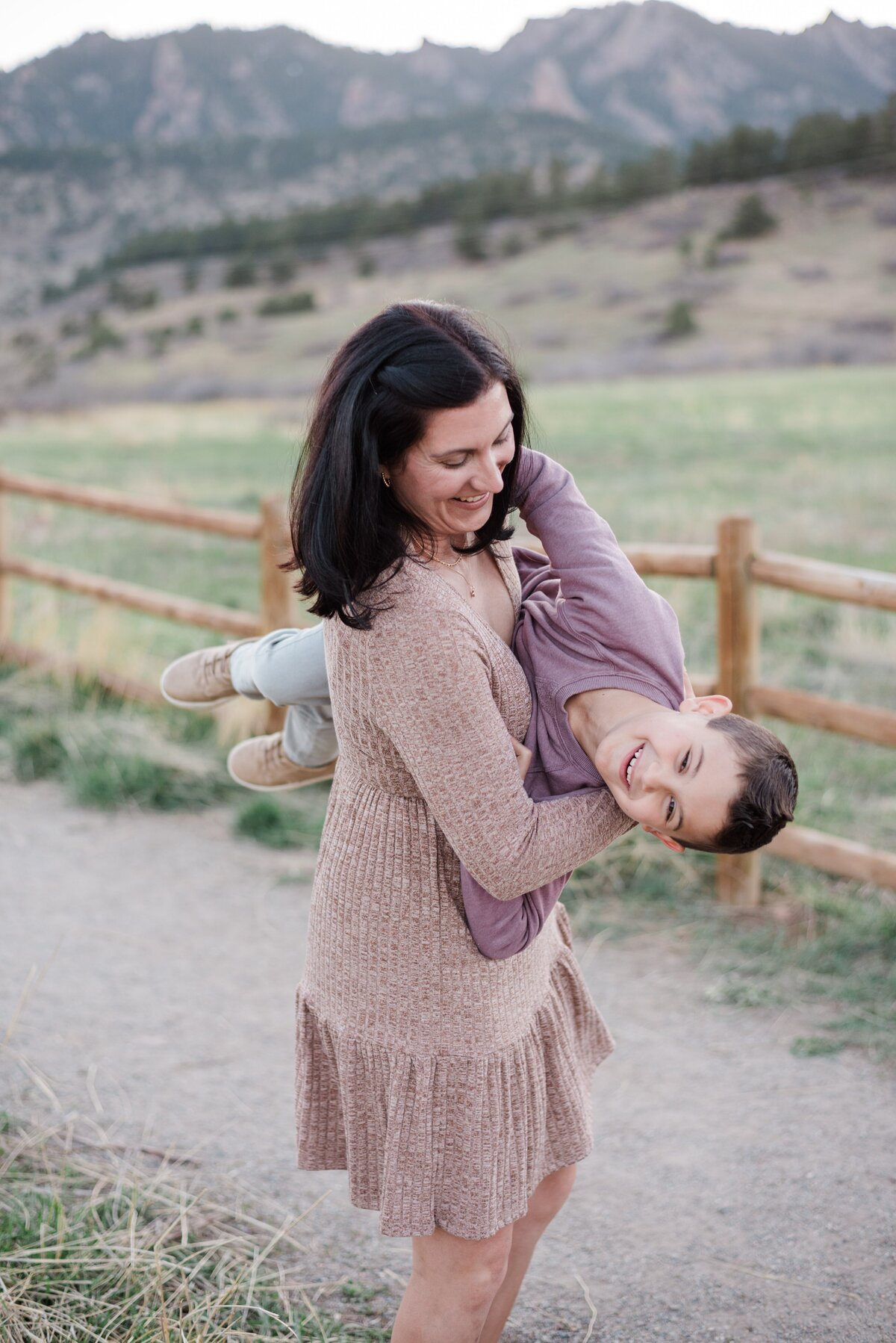 mom in a mauve long sleeve dress holds her young son sideways as he smiles towards denver family photographer and the mom smiles at her son