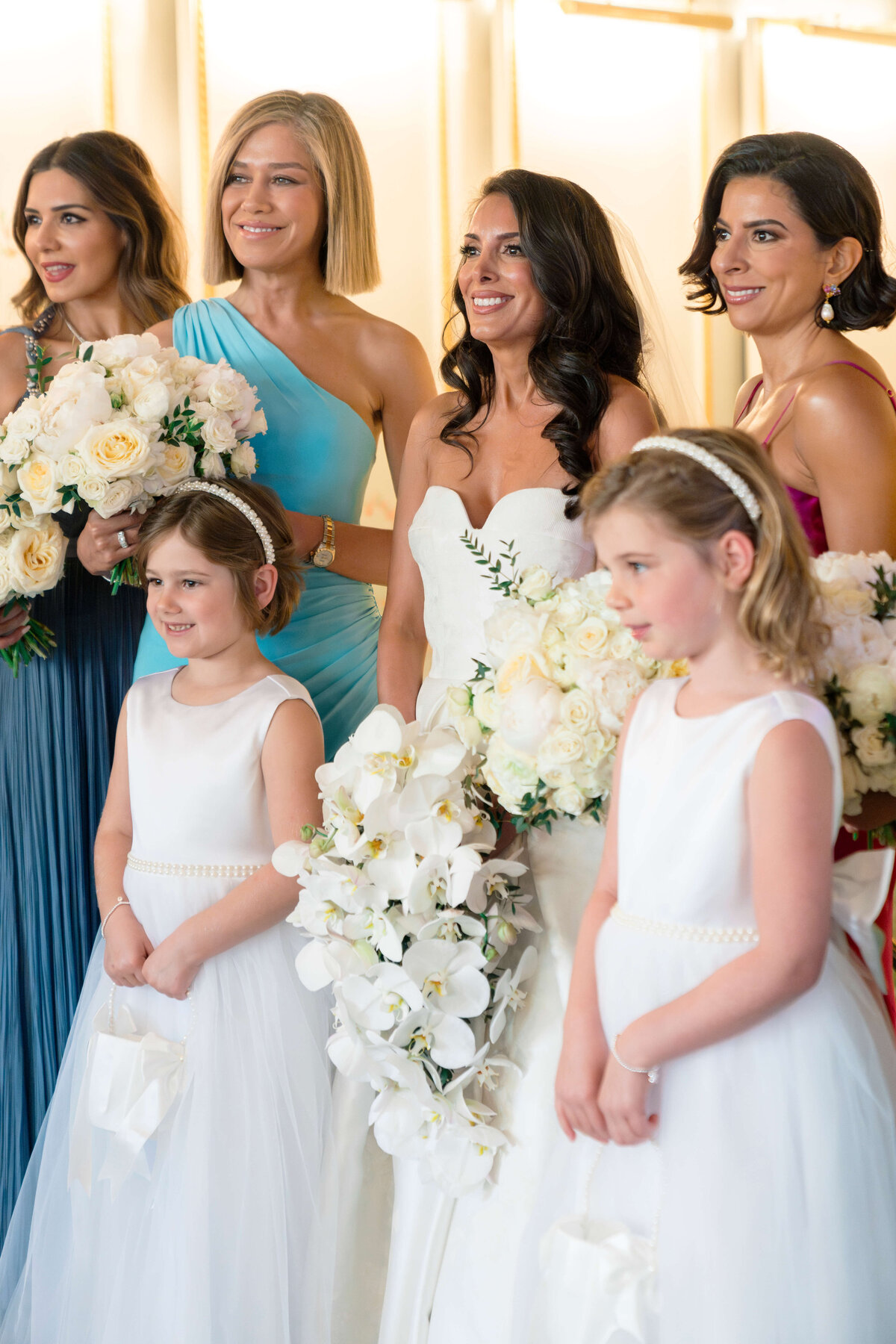 A group of women in elegant dresses and two young girls in white dresses stand together smiling. The women hold bouquets of white flowers, and the scene is a formal occasion.