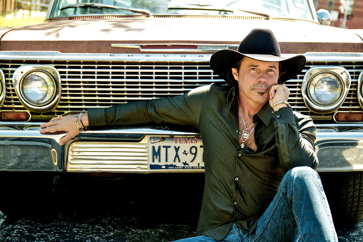 Country Music Photography Derryl Perry sitting on ground in front of grill of old car wearing big black cowboy hat hand on chin
