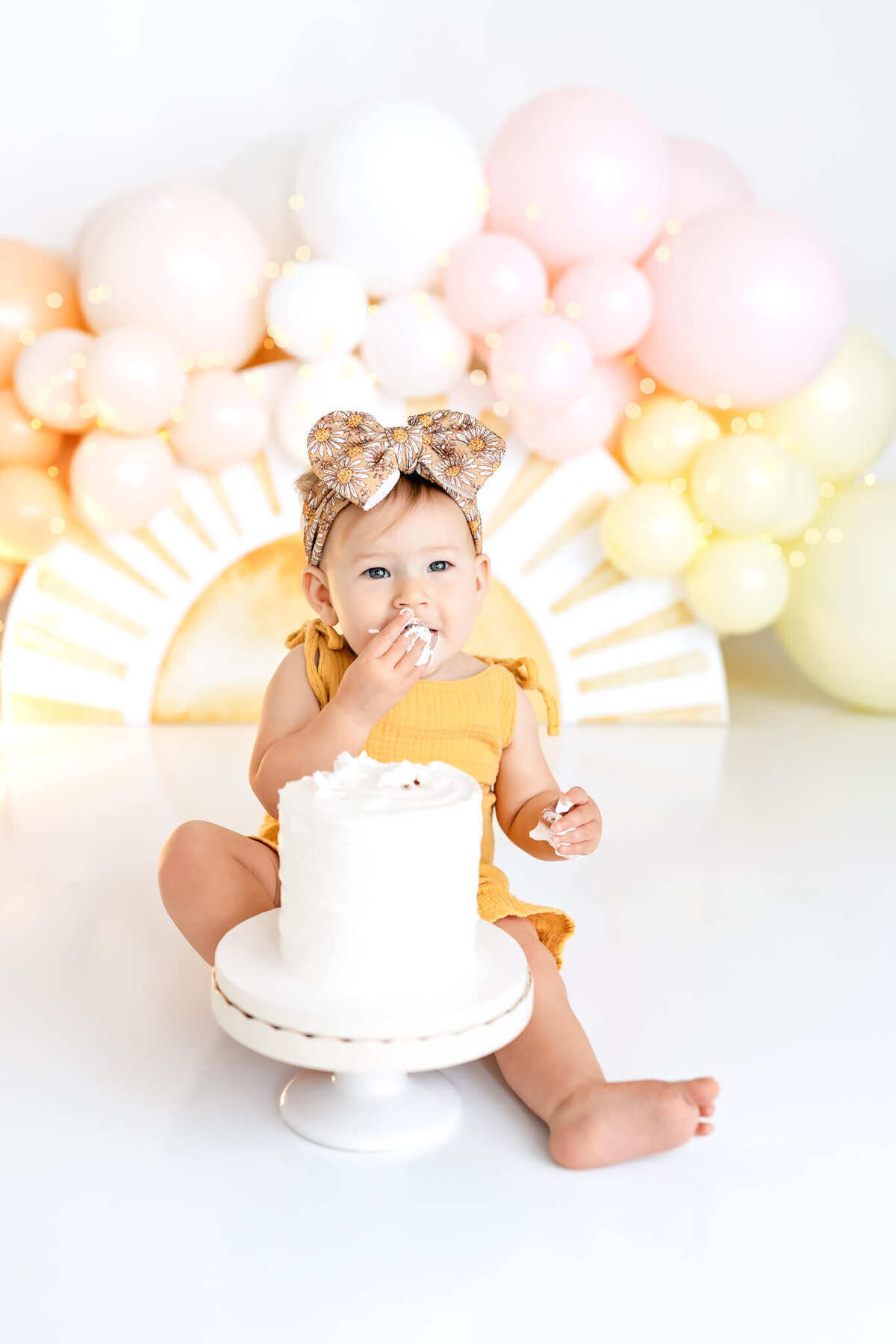 A baby wearing a large bow on her head sits on the floor, eating cake from a small white cake with her hands. The background has pastel-colored balloons arranged in an arch. The baby is dressed in a yellow outfit.