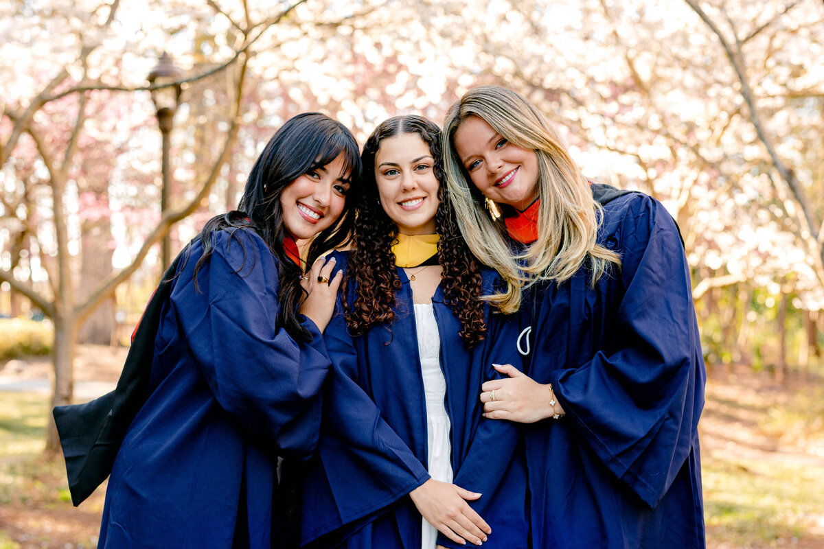 Group of three Duke graduates in gowns and hoods embracing and smiling in front of cherry blossoms.