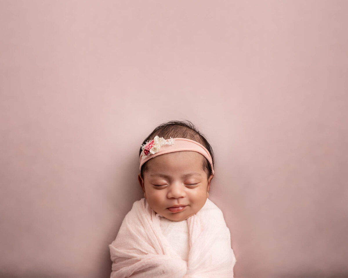 A newborn baby girl laying on a pink blanket and wrapped in pink smirks while having her photo taken during a session with an Asheville Newborn Photographer