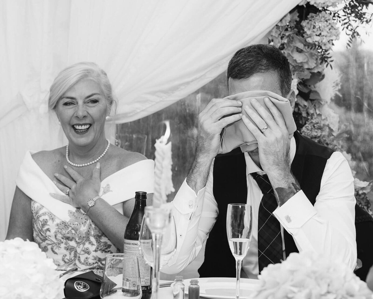 The mother of the bride laughs as the groom covers his face during the best man speech
