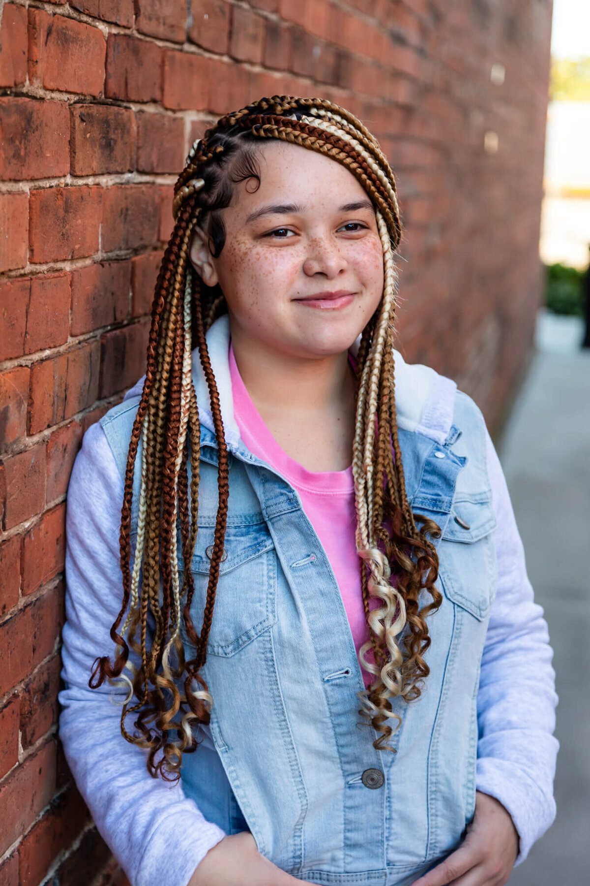 A lovely girl with braids wearing pink and light wash denim hoodie leaning on a brick wall. Captured by Springfield, MO senior photographer Dynae Levingston.