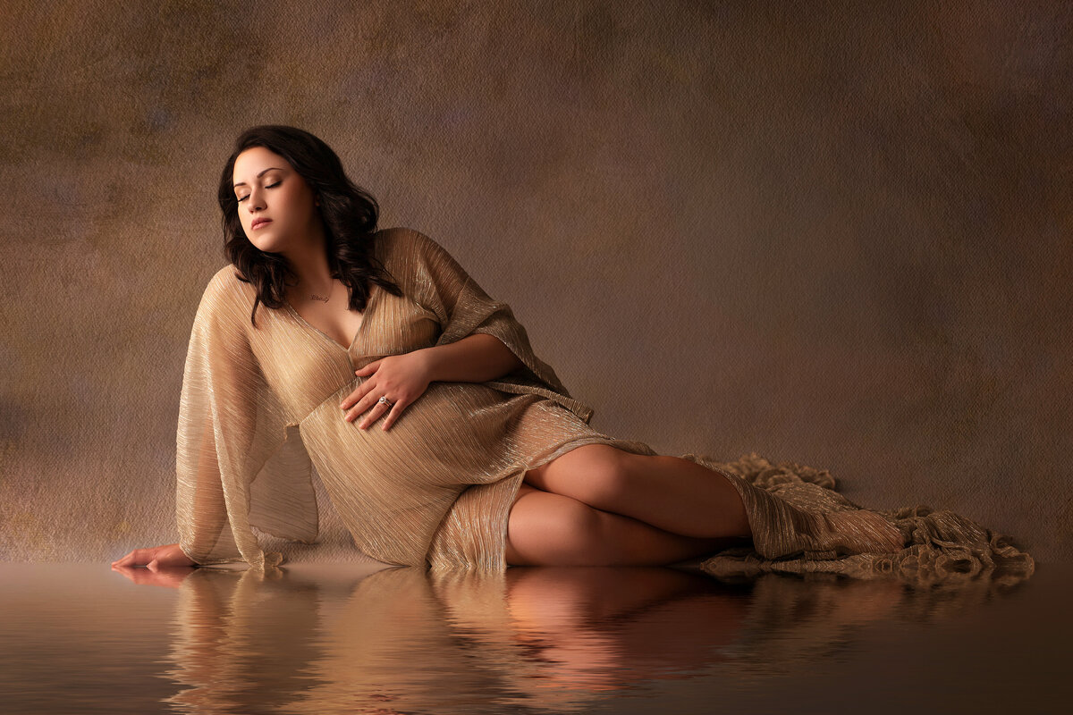 Pregnant woman in a soft, draped white gown cradling her belly while seated on a floor in a bright Waukesha studio, showcasing wardrobe options provided by the photographer.