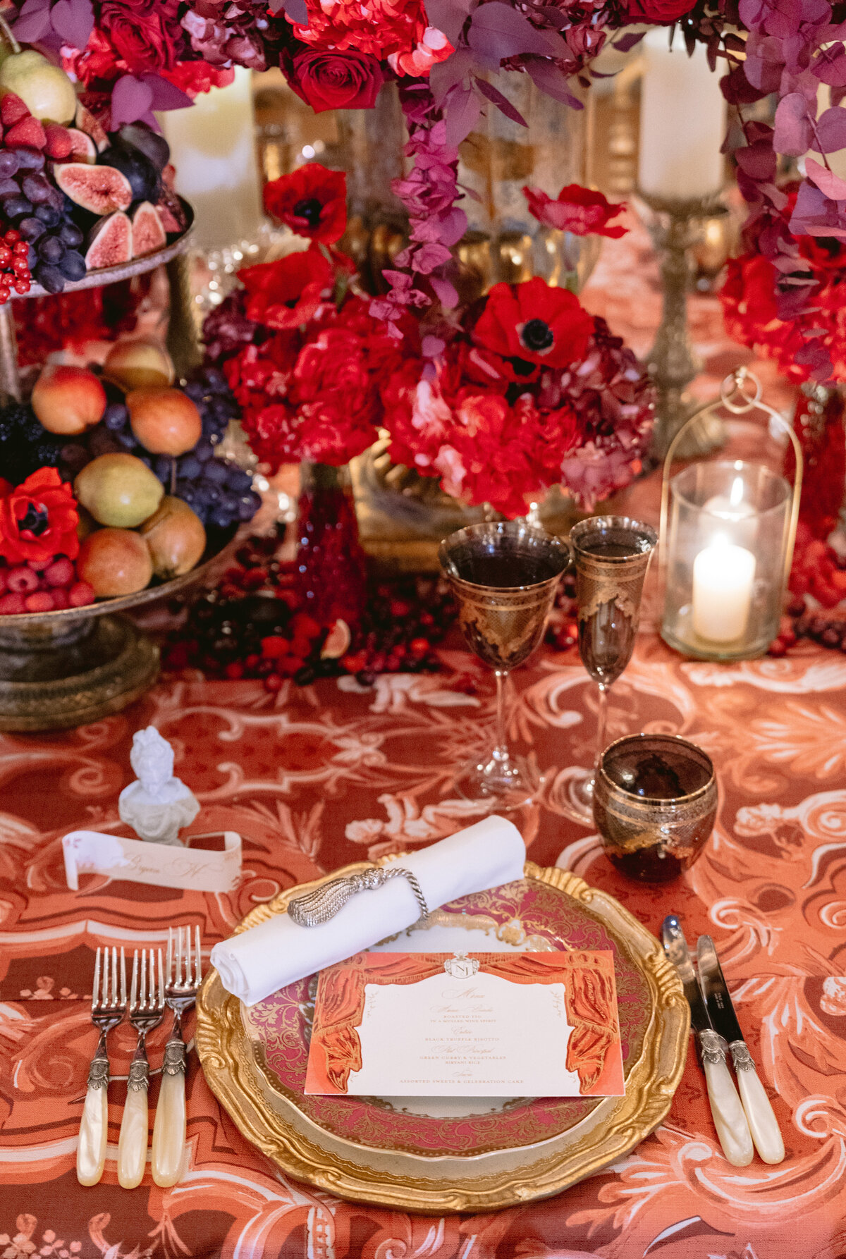 A lavishly set table features a gold-rimmed plate with a red and white menu, a neatly rolled napkin held by a jeweled ring, various cutlery, and ornate glasses. The centerpiece includes vibrant red flowers, fruits, and lit candles, all atop a red patterned tablecloth.