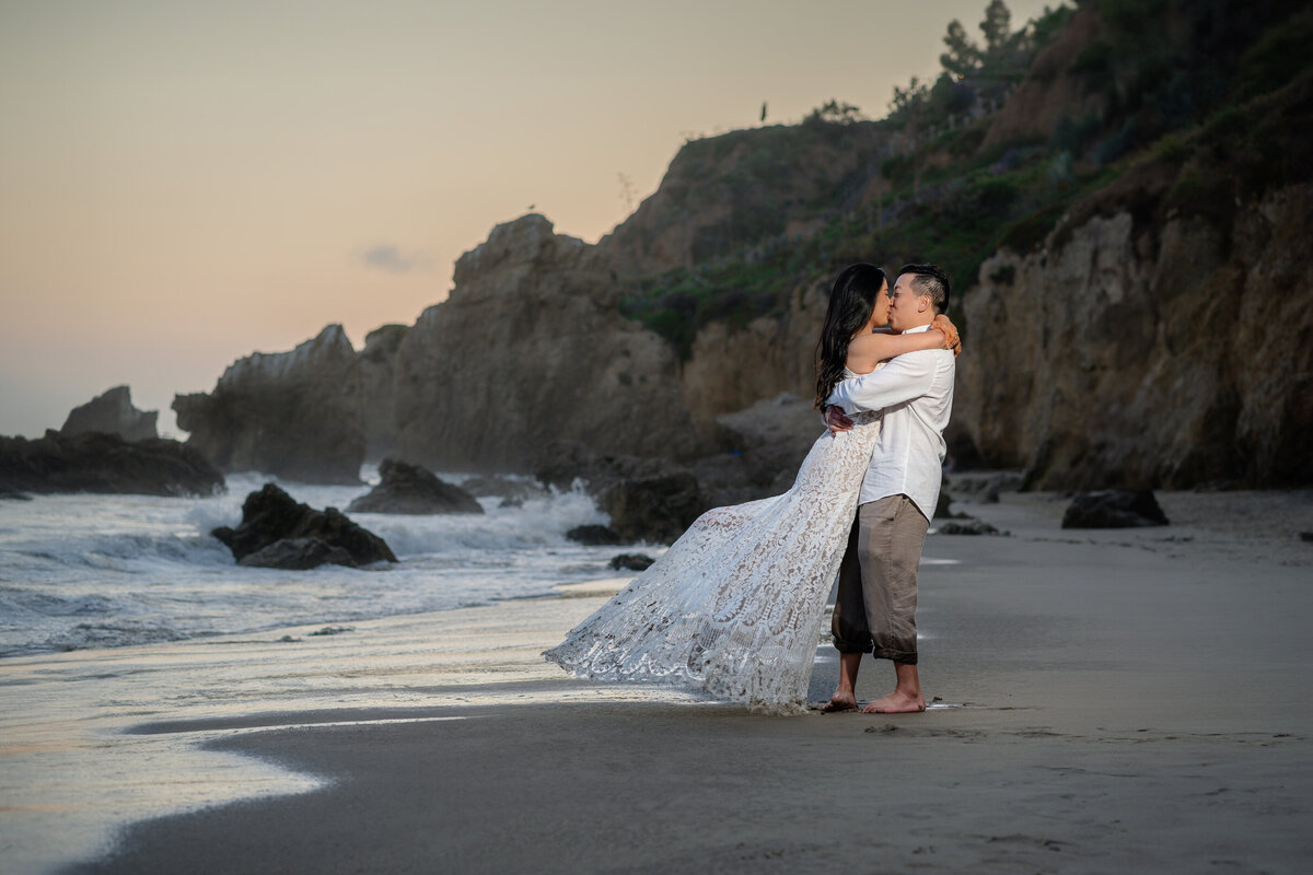 Asian couple eloping at El Matador State Beach in Malibu during low-tide sunset and kissing and lifting with rocky cliff beach.