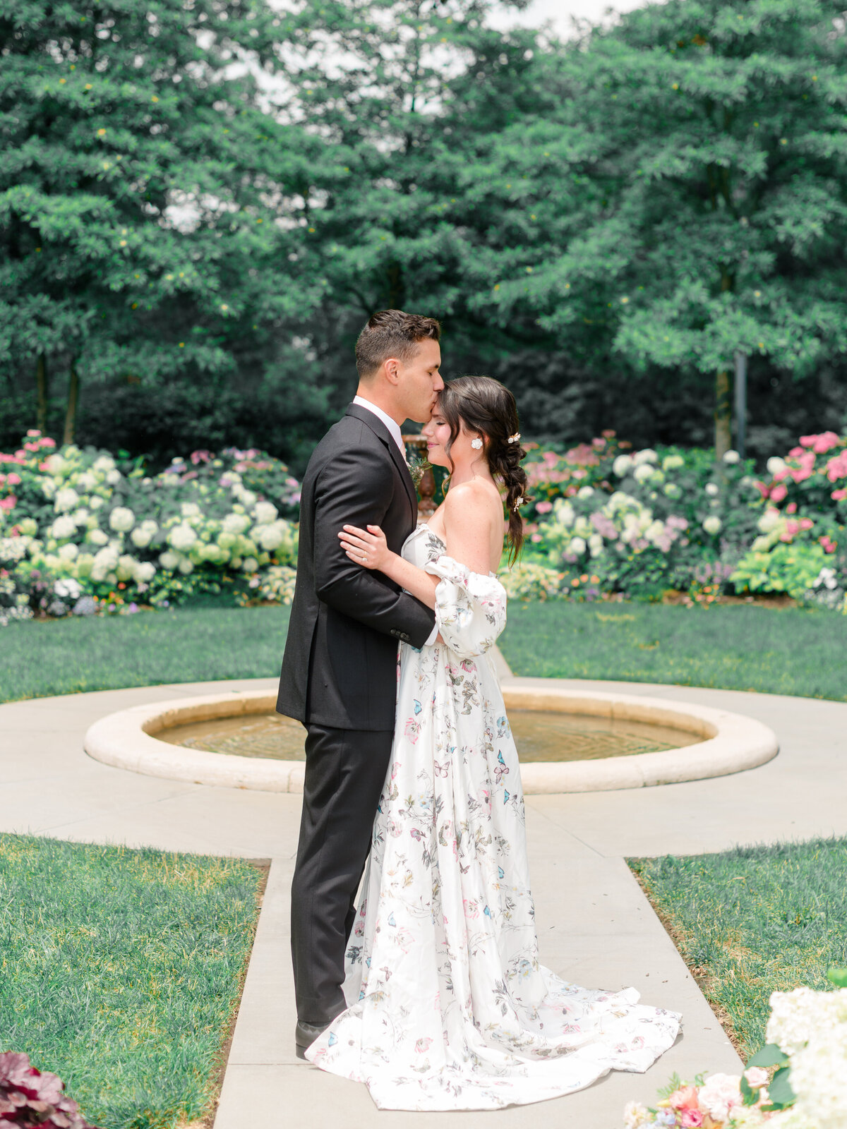 Romantic garden portrait of a bride and groom embracing in front of the fountain at Cairnwood Estate in Bryn Athyn, Pennsylvania. Captured by Elizabeth Kane Photography, a Philadelphia Fine Art Wedding Photographer, this timeless image highlights floral-filled grounds, classic East Coast elegance, and heartfelt editorial storytelling.
