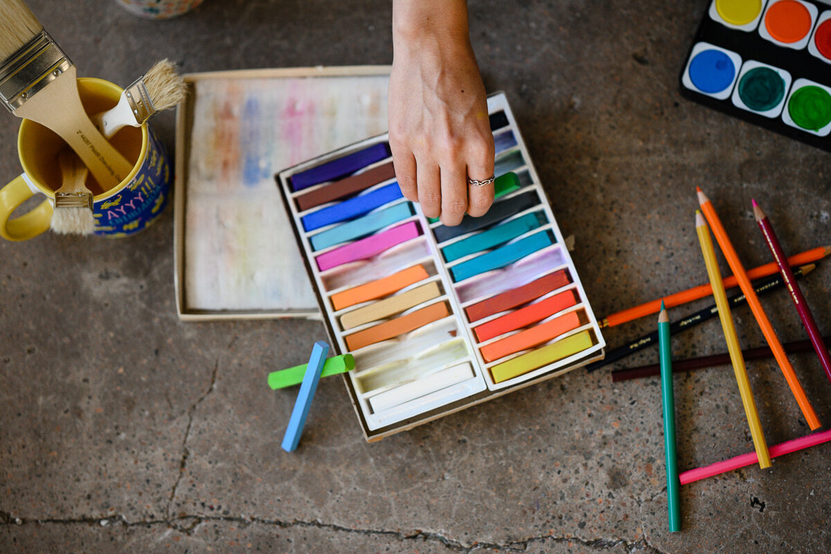 A stock image for an artist taken during her branding photos by a Denver brand photographer that is her reaching into a box of art supplies that are laid all over a concrete floor.