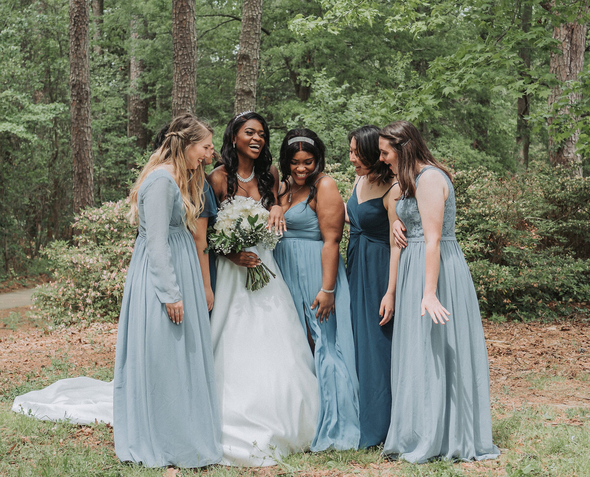 A joyful bride in her white wedding gown stands with her bridesmaids dressed in varying shades of blue, laughing and smiling together in a natural park setting in Virginia Beach, captured by Quinn Photography LLC.