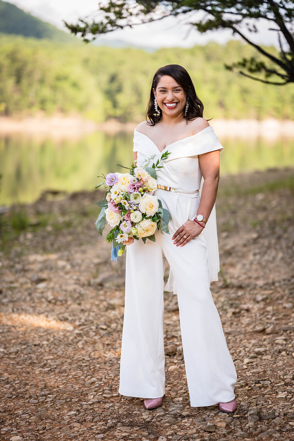 A bride wearing a jumpsuit and holding a bouquet of pastel flowers stands along the shores of Carvin’s Cove for a formal portrait on her elopement day.