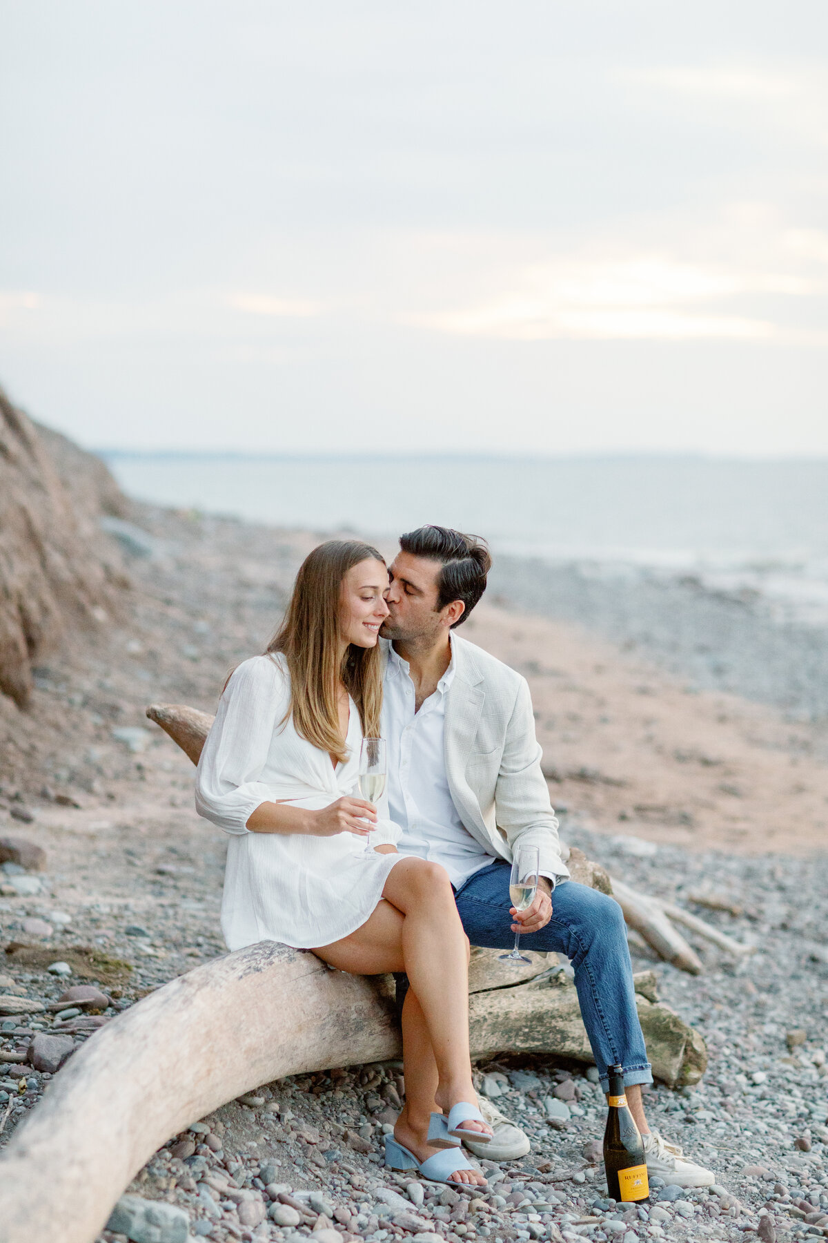 Couple sitting on driftwood and holding champagne glasses during their engagement session at Chimney Bluffs.
