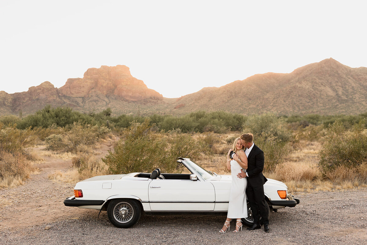 couple embracing while standing in front of a white classic vintage car in front of red mountain in mesa arizona