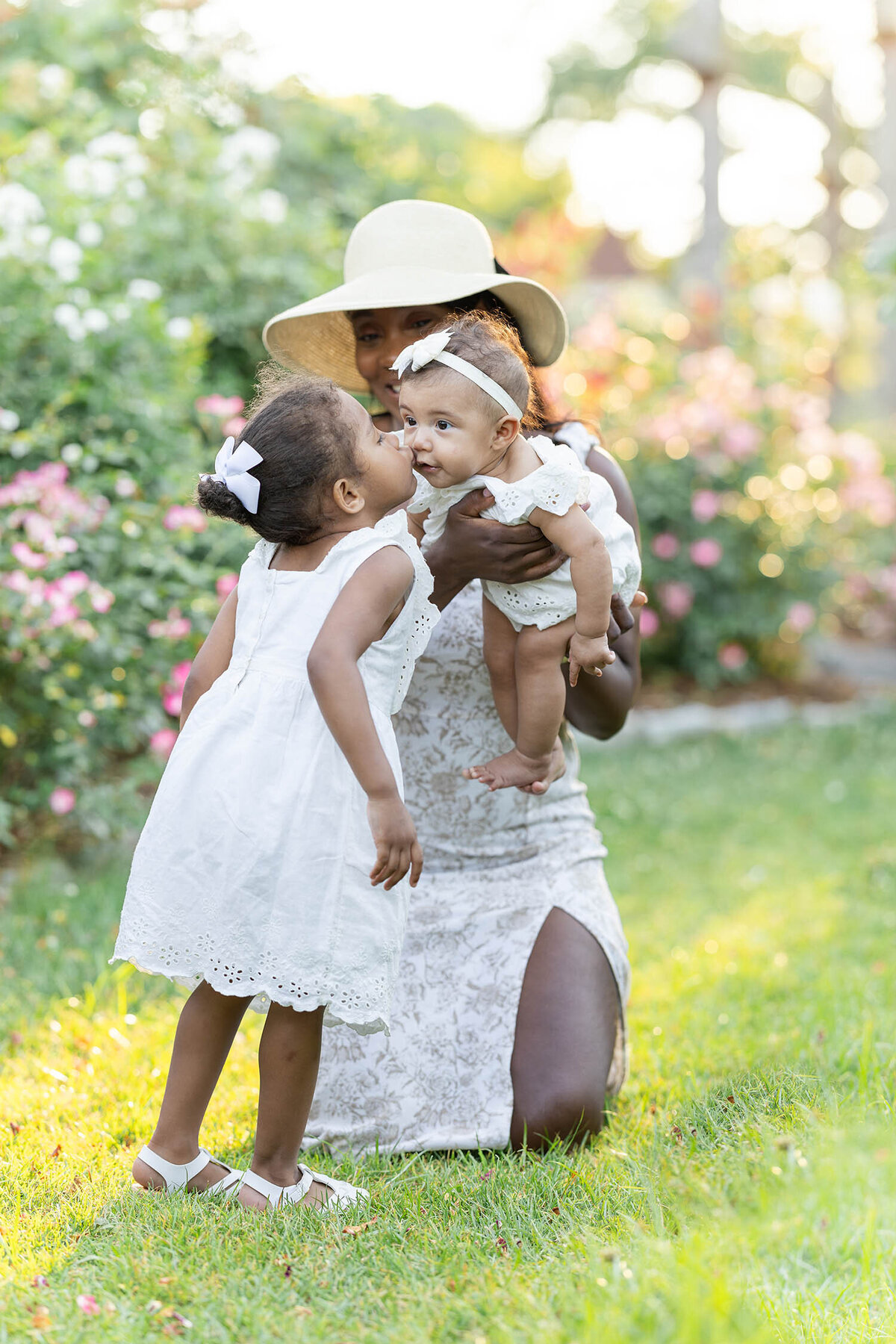Toddler girl gives baby sister a kiss as mom holds baby sister out towards toddler girl during their mommy and me session in a rose garden
