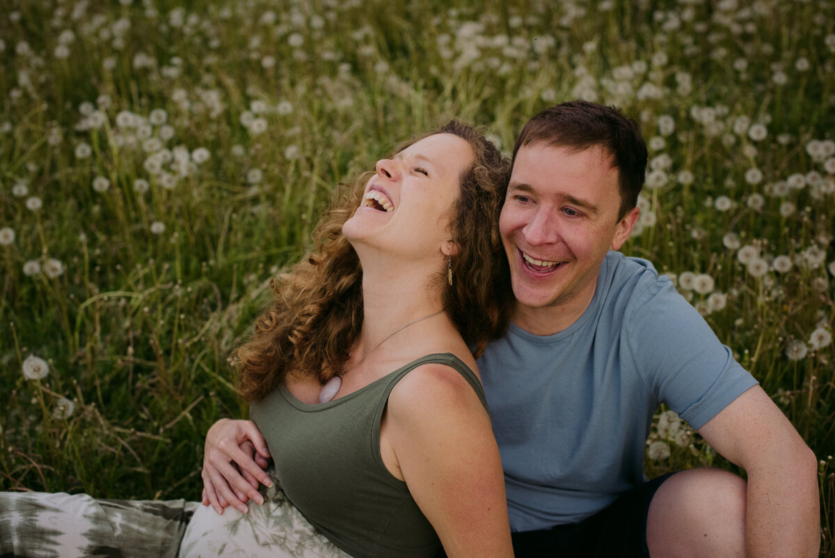 pregnant girl sitting with her husband in a field of dandelions laughing
