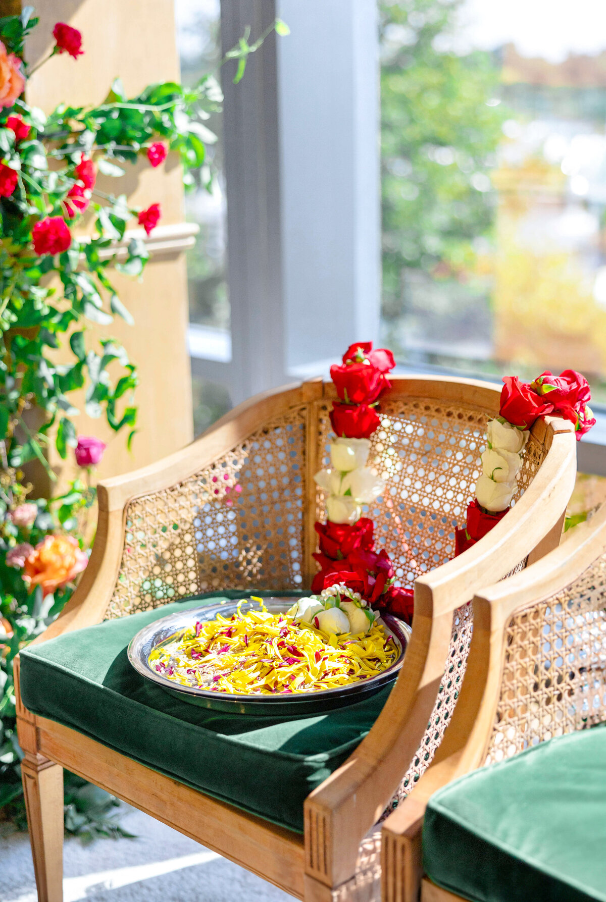 A wicker chair with a green cushion holds a plate of yellow flowers and two garlands made of red and white flowers. The background features a window and floral decorations, with sunlight streaming in.