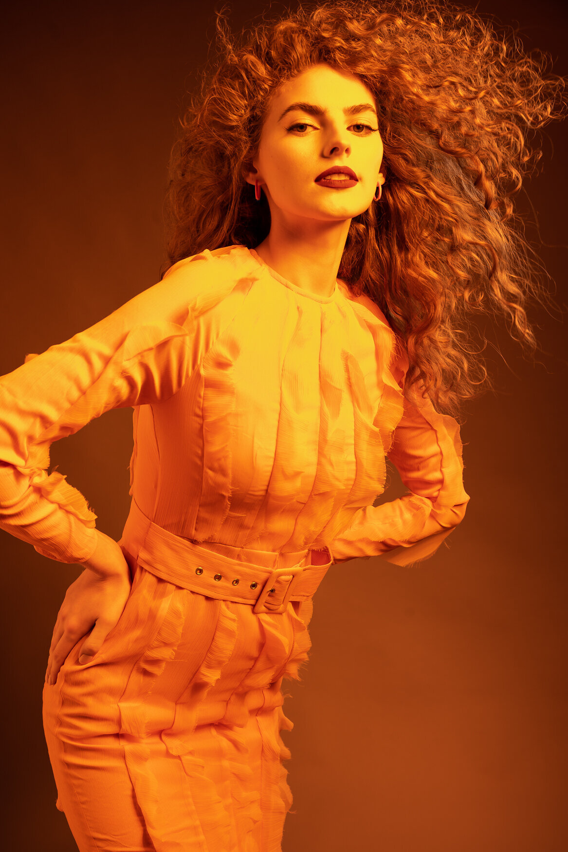 Portrait of a woman standing confidently with hands on her hips, drssed in an orange textured dress with a belted waist.