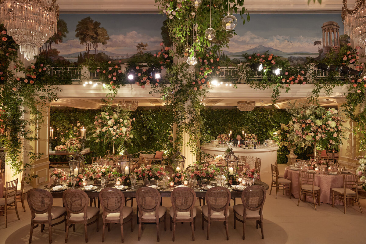 The photo was taken at The Willard Intercontinental in Washington D.C., one of the top wedding venues and hotels in DC and captured by photographer Shawn Connell. The event space featured elegantly set dining tables with floral centerpieces, surrounded by greenery and hanging lights. The room also had a balcony adorned with greenery and a scenic mural depicting a classical landscape above it. Additionally, Clayton Films served as the videographers for this event.