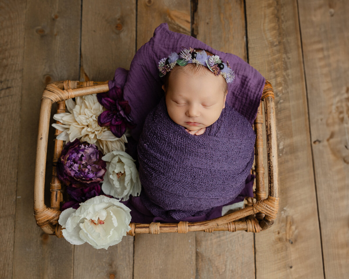 Purple-themed newborn portrait featuring a sleeping baby girl styled with soft fabrics and florals on a wooden floor. Captured by Inland Empire newborn photographer Kaitlyn Dawn.