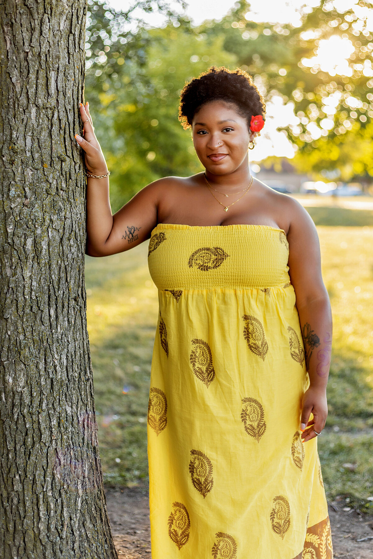 Woman in yellow halter sundress with red flower in her hair leaning on tree at sunset
